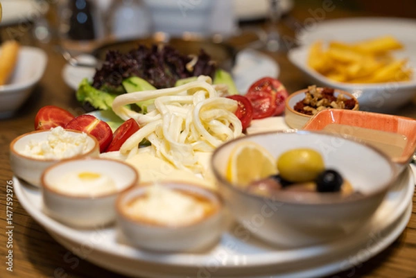 Fototapeta Close up view of a mixed breakfast plate with various foods, including cheese, jams, butter, honey, walnuts, tomatoes, and olives, on a restaurant table.