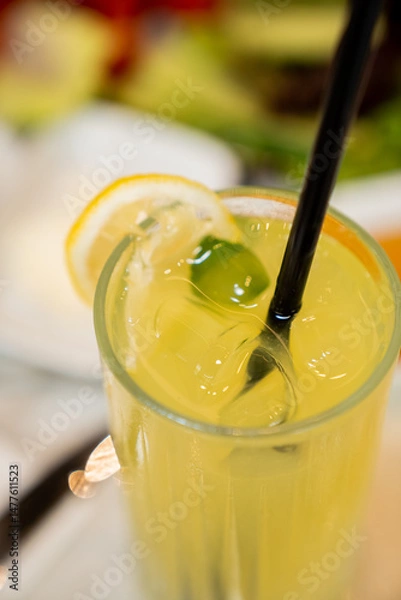 Fototapeta A glass of lemonade with ice, a lemon slice, fresh mint, and a straw is served on a restaurant table.