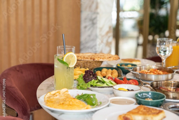 Fototapeta Close up view of a mixed breakfast table with various foods, including jams, cheese, fresh lemonade, butter, pastry, vegetables, olives, and an omelette, on a restaurant table.