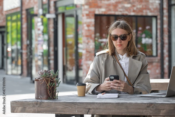 Obraz Young woman working using phone and laptop sitting on cafe veranda. Remote work, businesswoman