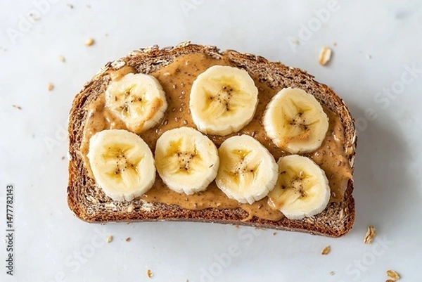 Fototapeta A slice of whole grain bread with a spread of almond butter, topped with banana slices, placed on a clean white surface