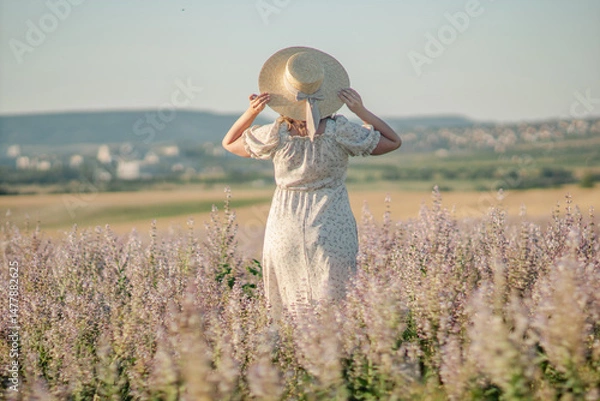 Obraz Woman Hat Field: Summer rural landscape; woman in white dress carries hat, walking through purple wildflowers for sun protection.