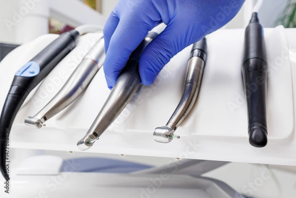 Obraz A gloved hand selects a dental handpiece from a set of tools on a white holder. The clinical setting features neatly arranged instruments and a clean background.
