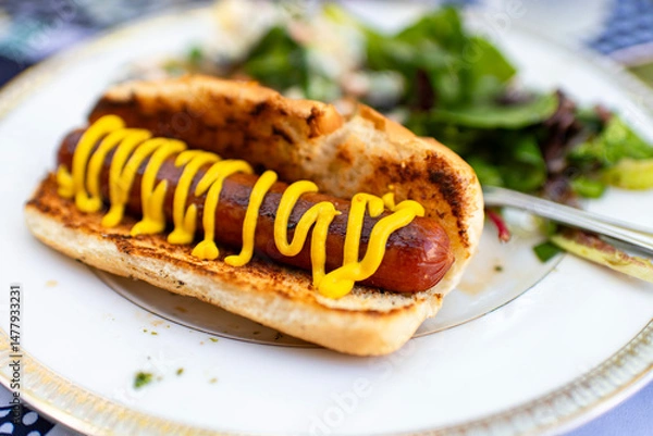 Obraz A hot dog with mustard on it on a plate with salad in the background 
