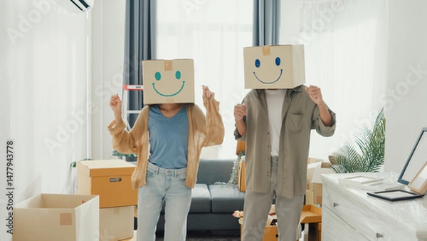 Fototapeta Happy Asia couple stand front of sofa couch used carton box made smiling face funny dance front of camera together in living room at new house. Husband and wife moving new house, Couple goal Concept.