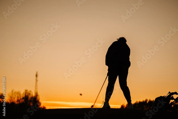 Obraz Silhouette of a female golfer hitting a ball at sunset, with club extended and golf bag nearby, against a vibrant orange sky on a peaceful course..