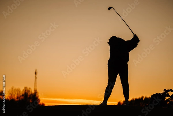 Obraz Silhouette of a female golfer finishing a full swing during sunset, with golf bag beside her and an orange sky in the background..