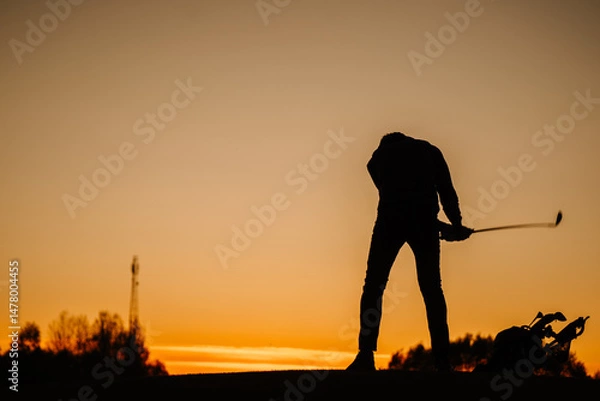 Obraz Silhouette of a golfer bending with club in hand at sunset, next to a golf bag, set against a glowing orange sky and distant trees..