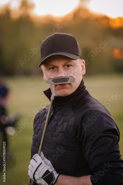 Obraz Male golfer reacts emotionally after a putt, raising his club and touching his cap on a golf course during sunset, surrounded by trees..