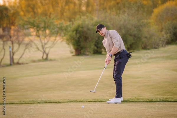 Obraz Male golfer mid-putt on a golf green during sunset, focused on hitting the ball with precision, surrounded by trees and open landscape..