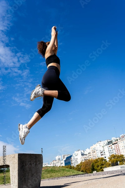 Fototapeta Young sporty woman jumping in urban park under blue sky