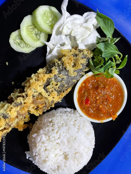 Fototapeta Pecel lele menu complete with fried catfish served with rice, cabbage leaves, sliced ​​cucumber and chili sauce ready to eat.