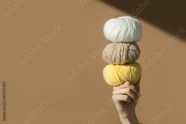 Fototapeta Woman's hands hold pyramid of threads wrapped in balls of yellow, white and brown colors on her finger. Beige background with copy space and rays of sunlight. Mockup for advertising