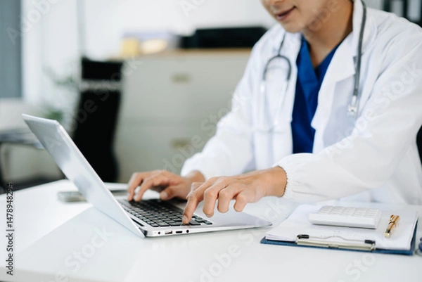 Fototapeta Doctor using laptop in a medical office with stethoscope and clipboard. Concept of digital healthcare, diagnostics, and patient data