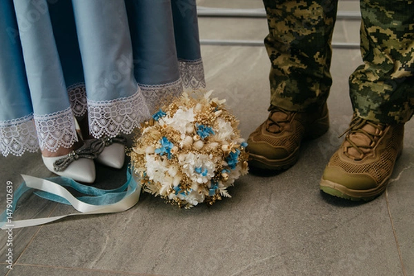 Obraz Between the legs of the bride in a blue dress and the groom, who is a military man from Ukraine, lies a wedding bouquet of colorful flowers