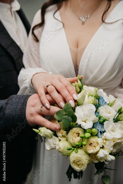 Obraz a couple in love on their wedding day pose for the photographer hugging each other and placing their hands with gold wedding rings on their fingers, on a wedding bouquet of white roses
