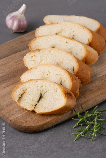 Fototapeta Freshly sliced bread with garlic and herbs displayed on a wooden cutting board on grey table