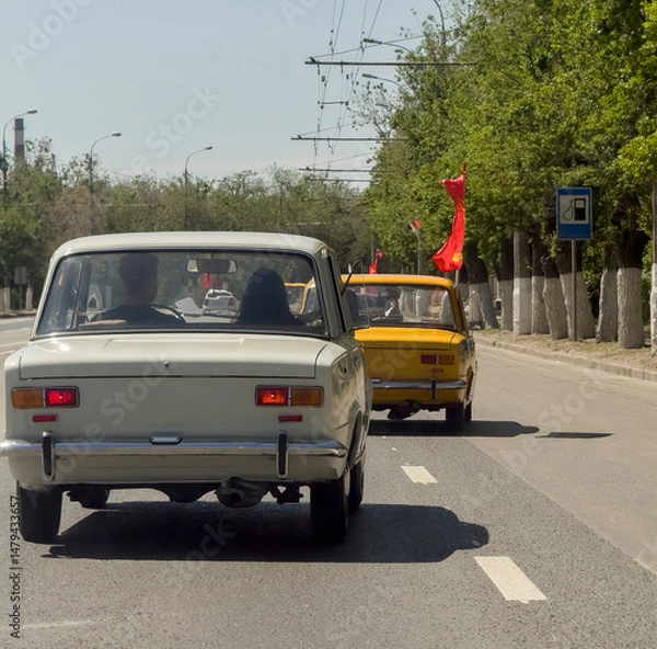 Obraz A vibrant retro car is driving along a picturesque road, with another classic vehicle seen in the background on a sunny day.