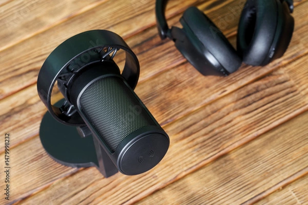 Fototapeta Top-down shot of a black studio microphone resting on a wooden desk with headphones in the background, ideal for podcast production and live streaming visuals. Photo, selective focus