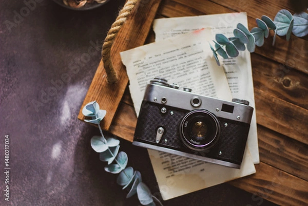 Fototapeta Retro camera on old book and eucalyptus branch on wooden background, flat lay. Vintage style