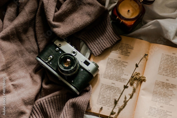 Fototapeta Vintage film camera with an old book wrapped in a warm knitted sweater and an extinguished candle next to it. View from above on a flat surface.