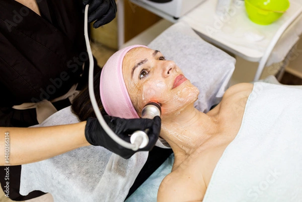 Fototapeta A woman between 50 and 60 years old undergoes an electro-aesthetic radiofrequency therapy. Concept of radiofrequency in older women.