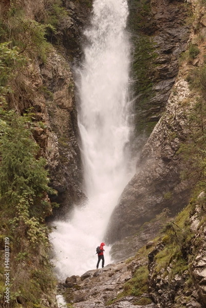 Obraz A lone hiker in a red helmet and backpack stands at the base of a powerful waterfall cascading down rugged cliffs. The towering flow of water and dramatic rocky landscape evoke awe.