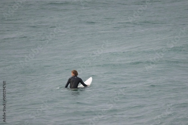 Obraz A surfer in a black wetsuit rides a breaking wave near the shoreline on a cloudy day. The powerful movement of the sea and the surfer’s focus capture the spirit of adventure and balance