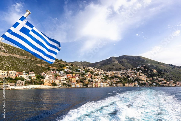 Fototapeta Colorful buildings on a hillside in the idyllic town of Symi by the Aegean Sea on Symi (Simi, Syme) island in Greece on a sunny day. Greek flag is in the foreground.
