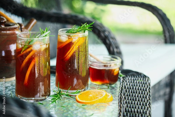 Fototapeta Cold ice herbal tea with orange slices and rosemary stem leaves in  tall glasses. Selective focus on second glass with blurred foreground and background..