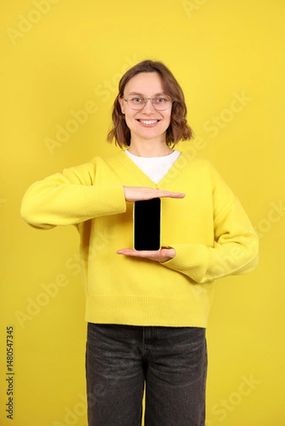 Obraz Mobile App promotion. Cheerful young woman showing black empty smartphone screen posing over yellow background. Cellphone display mock up. Online service marketing, digital product advertisement