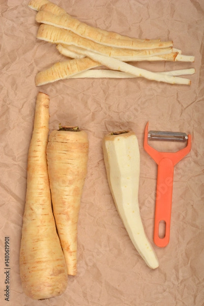 Fototapeta Peeled and unpeeled parsnips lie on a brown paper surface next to an orange vegetable peeler
