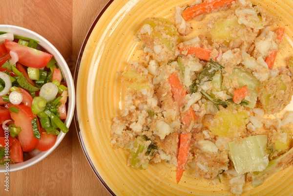Fototapeta Cooked vegetables with saladA top-down view shows a yellow plate with cooked carrots, cauliflower, and potatoes with breadcrumbs, next to a bowl of tomato and green onion salad on a wooden surface