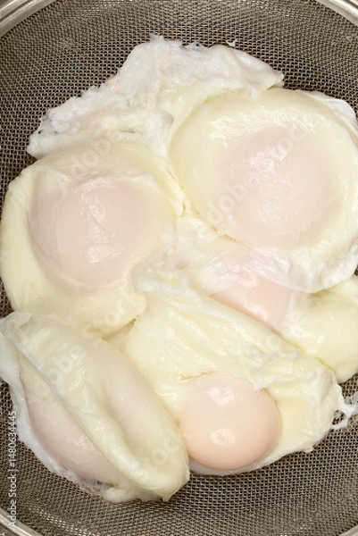 Fototapeta A close-up overhead shot shows several poached eggs with soft yolks and cooked whites resting in a metal sieve