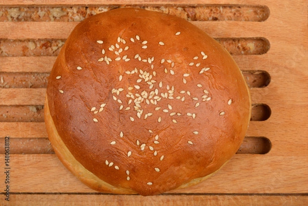 Fototapeta A single, golden brown homemade burger bun sprinkled with sesame seeds rests on a wooden cutting board