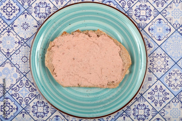 Fototapeta A slice of bread generously spread with light pink pate sits on a blue and white patterned plate