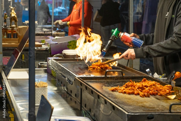Obraz Street Food Vendor with Dynamic Energy Cooking Spicy Chicken Using Torch Flame at Night Market Stall, Seoul, South Korea, Myeongdong Market