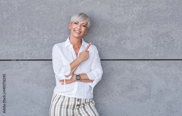 Fototapeta Presentation. Woman pointing to blank space - copy space, middle age woman presenting your product, against gray background - wall. Promoter shows space for your text