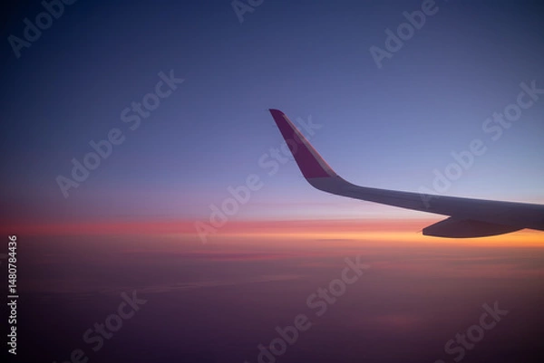 Obraz An airplane wing is captured against a stunning twilight sky filled with a gradient of blue purple orange and pink, symbolizing air travel and the serene beauty of a sunset from above the clouds.