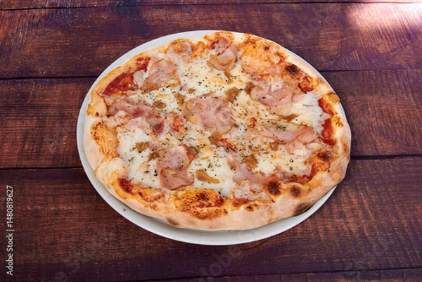 Obraz A pizza with ham and cheese on a white plate. The pizza is sitting on a wooden table