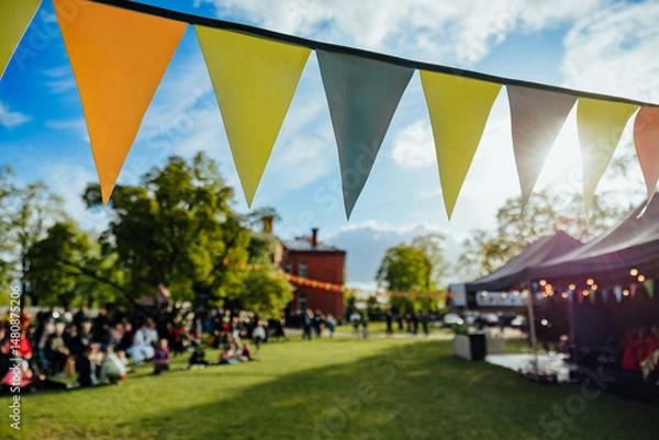 Fototapeta A lively outdoor festival scene with colorful bunting flags strung across a sunlit park, where people gather and enjoy a communal celebration under clear skies