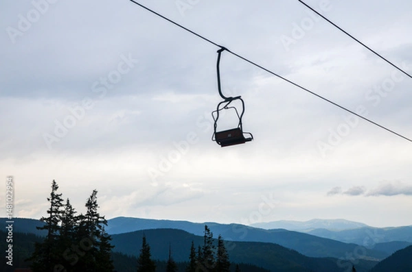 Fototapeta Single ski chairlift hangs suspended against a cloudy sky, with forested mountains in the background creating a peaceful and isolated atmosphere