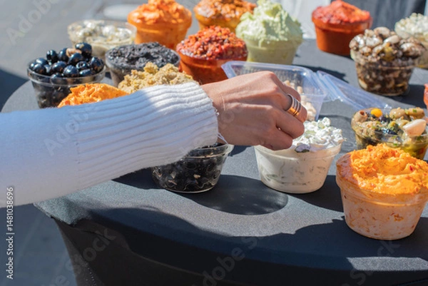 Fototapeta Woman's hand picking up a sample of fresh tzatziki sauce from a variety of dips and spreads displayed on a table at an outdoor food market, offering a colorful and tempting selection