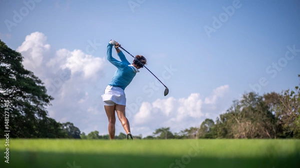 Fototapeta Professional female golfer lush green golf course, mid-swing after hitting a shot through after driving the ball down the fairway trees and distant sand bunkers, typical of a competitive setting.  