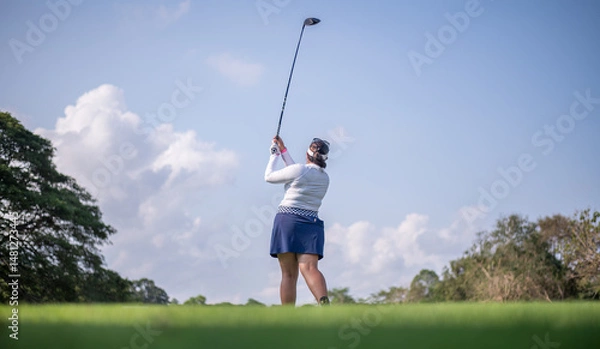 Fototapeta Professional female golfer lush green golf course, mid-swing after hitting a shot through after driving the ball down the fairway trees and distant sand bunkers, typical of a competitive setting.  