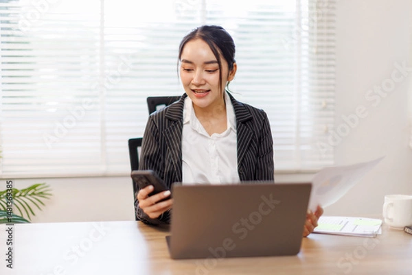 Obraz Happy asian woman works at home and uses a smart phone and a notebook laptop computer in the home office. Woman working on desk with using calculator, financial planning finance accounting.
