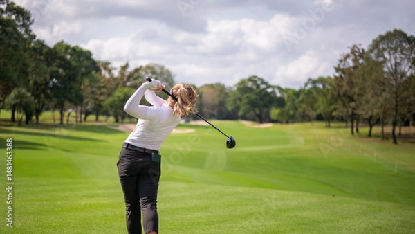 Fototapeta Professional female golfer lush green golf course, mid-swing after hitting a shot through after driving the ball down the fairway trees and distant sand bunkers, typical of a competitive setting.  