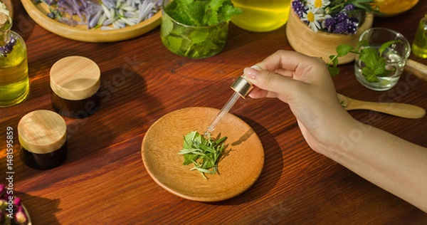Fototapeta A few dried rosemary leaves are placed on a wooden plate. A hand in the air is holding a dropper bottle and pouring drops of essential oil. Surrounding it are amber jars and other herbal medicine.