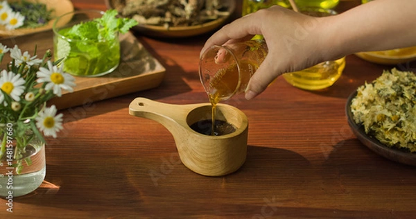 Fototapeta A hand is holding a beaker containing a yellow liquid and pouring it into a wooden cup with a handle. Inside the wooden cup are aromatic particles, mixed to form a brown medicinal mixture.