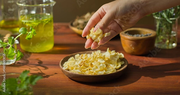 Fototapeta Dried petals are placed on a wooden plate, displayed on a brown table. A hand is holding the flowers and dropping them onto the plate below. Surrounding them are essential oil bottles and herbs.
